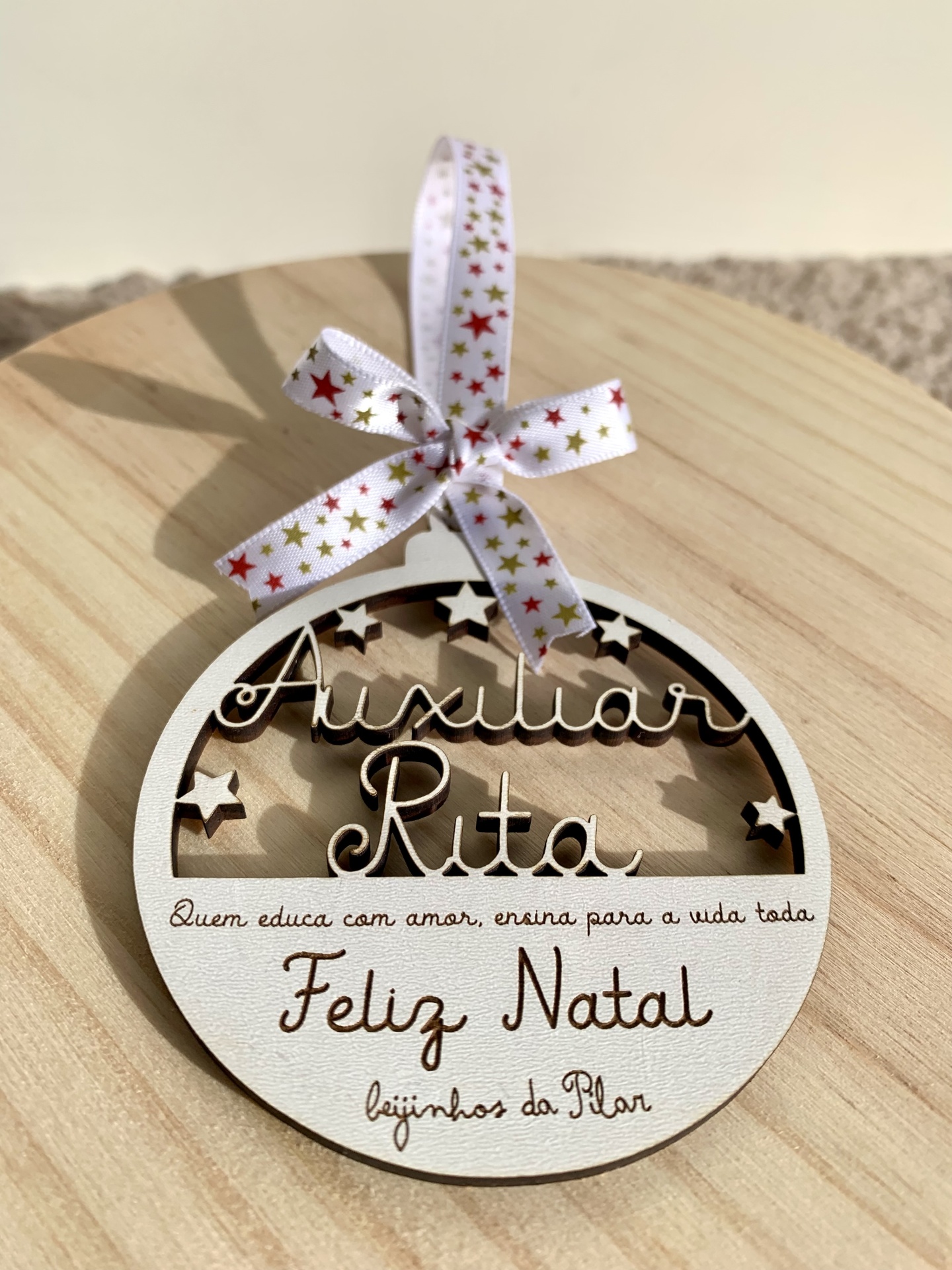 Enfeite de Natal redondo em madeira clara com texto e estrelas recortadas, pendurado em laço decorativo