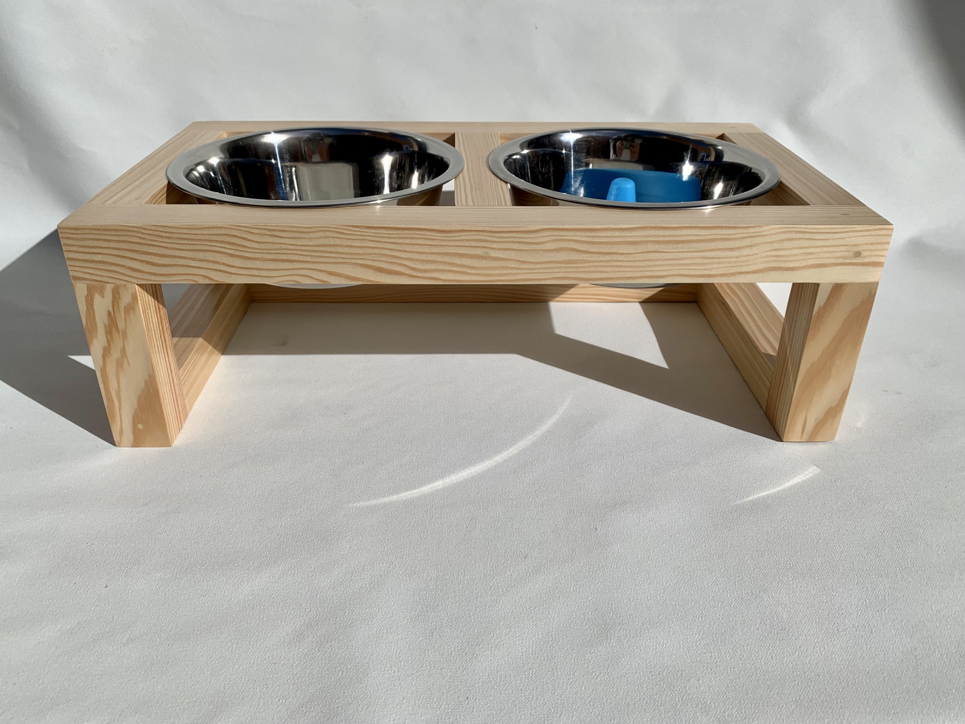 Comedouro duplo em madeira clara com duas taças metálicas, uma azul, sobre fundo branco