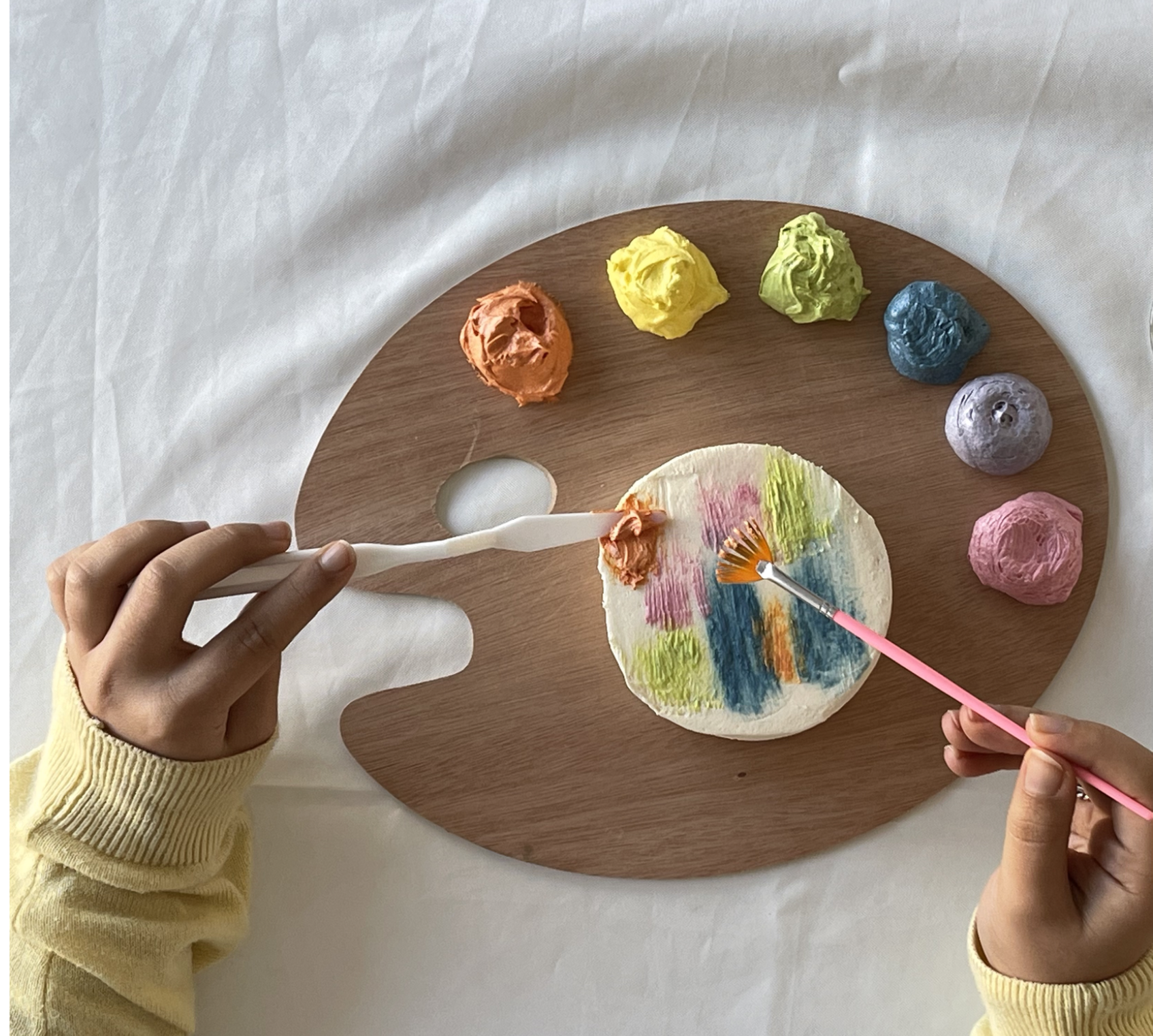 Paleta de madeira com tintas coloridas e mãos a pintar com pincel e espátula em fundo branco.