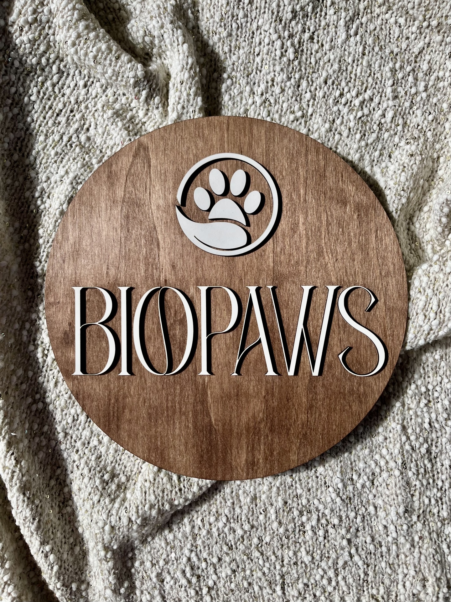 Placa redonda de madeira com logótipo e texto BIOPAWS sobre manta bege claro