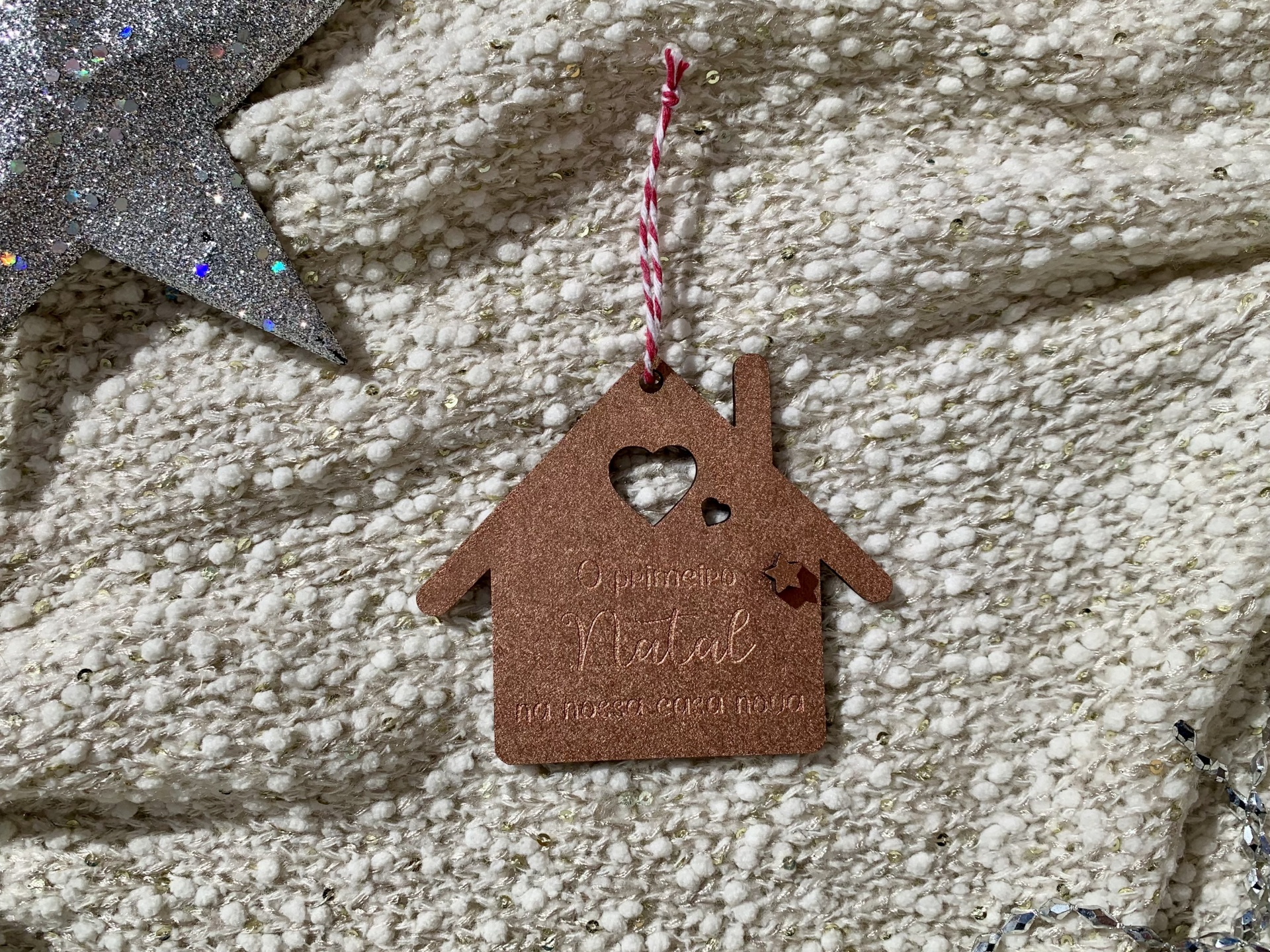 Enfeite decorativo em forma de casa castanho com cordão pendurado sobre tecido felpudo bege