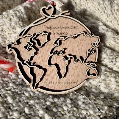 Ornamento redondo em madeira com mapa mundi recortado e texto gravado, pendurado sobre tecido cinzento com brilhos