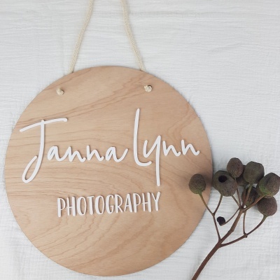 Placa redonda de madeira clara com texto branco pendurada numa parede branca e ramo seco ao lado