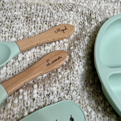 Talheres infantis de silicone verde água com cabos de madeira gravados com os nomes Miguel e Leonor ao lado de prato dividido verde água sobre tecido branco com detalhes dourados