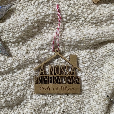 Enfeite pendente de madeira em forma de casa com texto e nomes, sobre tecido bege texturado