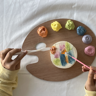 Paleta de madeira com tintas coloridas e mãos a pintar com pincel e espátula em fundo branco.