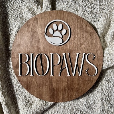 Placa redonda de madeira com logótipo e texto BIOPAWS sobre manta bege claro