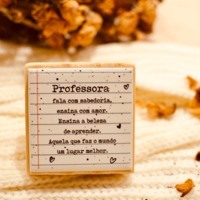 Peça de madeira com mensagem para professora, texto preto em fundo branco pautado, sobre tecido branco e elementos castanhos ao fundo.