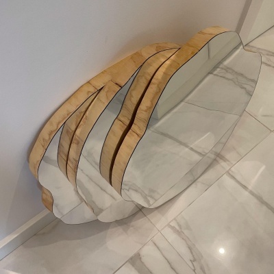 Espelho com moldura de madeira clara e forma irregular sobre chão de mármore branco