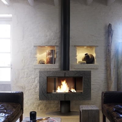 Lareira de ferro fundido cinza com padrão geométrico a preto e tubo de exaustão preto numa parede branca de pedra com nichos iluminados e mobília rústica