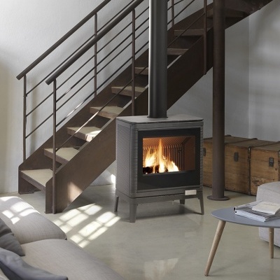 Lareira em ferro fundido cinzento com vidro frontal aceso instalada junto a escada metálica castanha em sala clara com sofá e mesa baixa