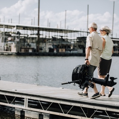 Casal de idosos a caminhar num cais junto à água com barcos ancorados ao fundo