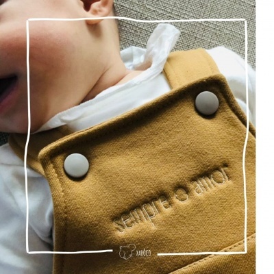 Suspensório de bebé castanho claro com bordado e botões, usado por criança com camisola branca