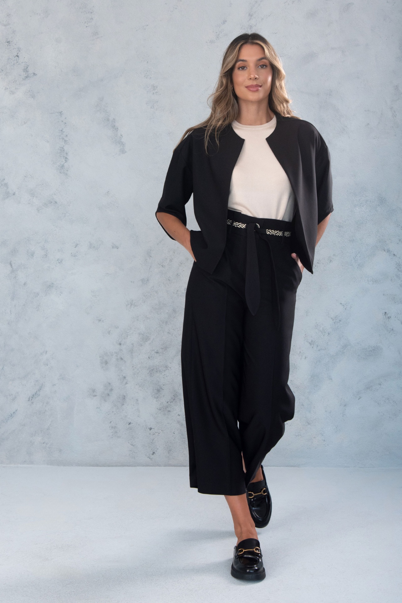 Calça fluida com cintura subida e cinto 200763 Mulher com casaco preto de manga curta, blusa branca, calças largas pretas e sapatos pretos