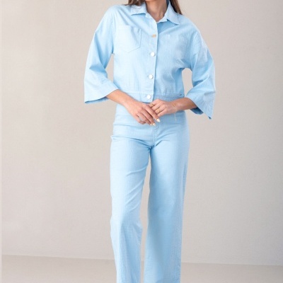 Mulher vestindo conjunto azul claro com camisa de botão mangas 3/4 e calças retas, sandálias brancas em fundo neutro