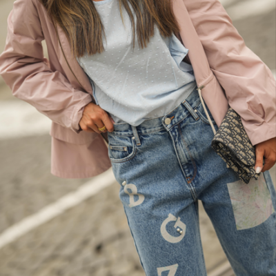 Mulher com calças de ganga azuis estampadas, t-shirt azul claro e blazer rosa claro, segurando carteira preta com padrão Dior