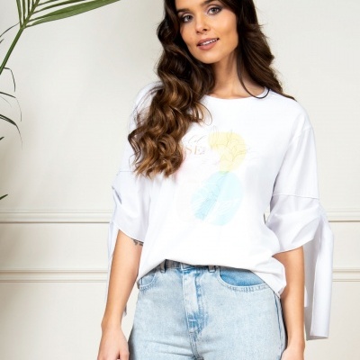 Mulher com camisola branca estampada e calças de ganga azuis em ambiente interior com planta verde