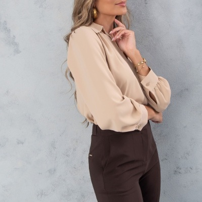 Mulher com blusa bege de manga comprida e calças castanhas contra parede cinza