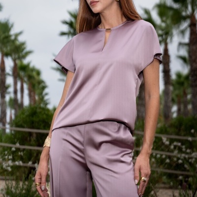 Mulher vestindo conjunto acetinado lavanda com blusa de manga curta e calças largas ao ar livre com palmeiras ao fundo