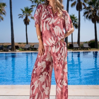 Mulher com conjunto rosa e branco de camisa e calças largas junto a piscina e palmeiras