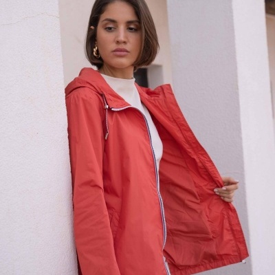 Casaco impermeável coral com fecho branco e capuz, mulher junto a parede branca