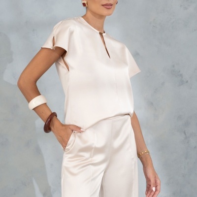 Conjunto feminino bege claro acetinado com blusa e calças largas, acessórios e fundo cinzento.