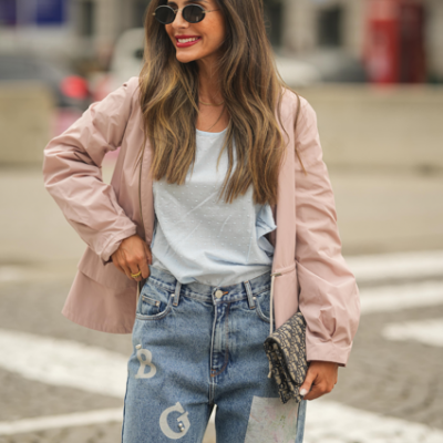 Mulher com casaco rosa, calças de ganga azuis com letras brancas, óculos de sol redondos e carteira padrão animal print