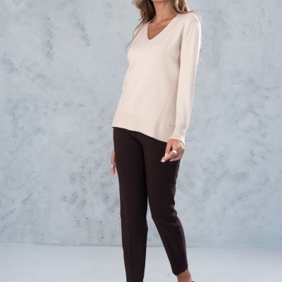 Mulher com camisola bege, calças castanhas e sandálias de salto em fundo cinza claro