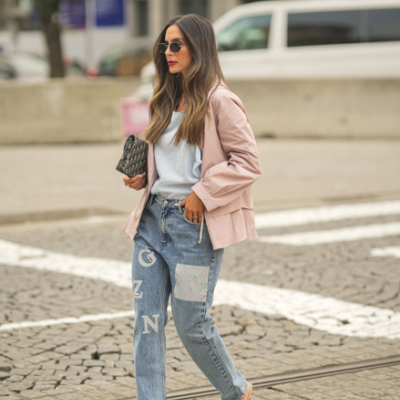 Mulher com casaco rosa, t-shirt branca, calças de ganga decoradas e sandálias nude atravessando a rua.