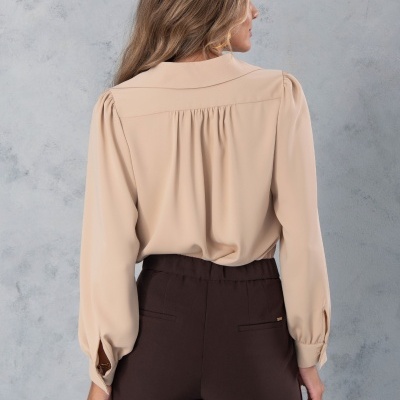 Mulher vista de costas com camisa bege e calças castanhas em fundo cinzento.