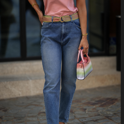 Mulher com t-shirt rosa, jeans azuis, cinto bege, sandálias verdes e mala multicolor pastel, em pavimento de pedra e degraus de granito