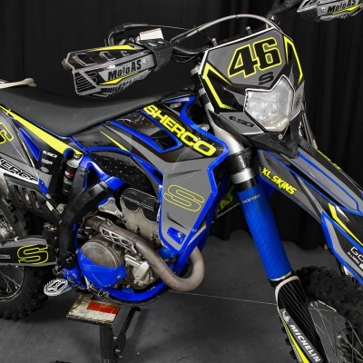 Motociclo de cross Sherco azul e preto com detalhes em amarelo sobre fundo escuro.