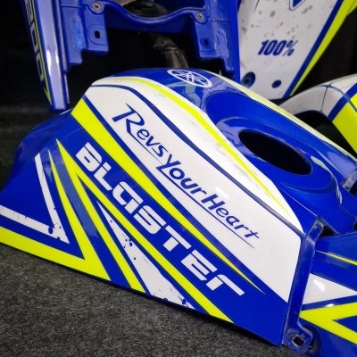 Peça para moto azul, branco e amarelo com texto 'Revs Your Heart' e 'BLASTER'.