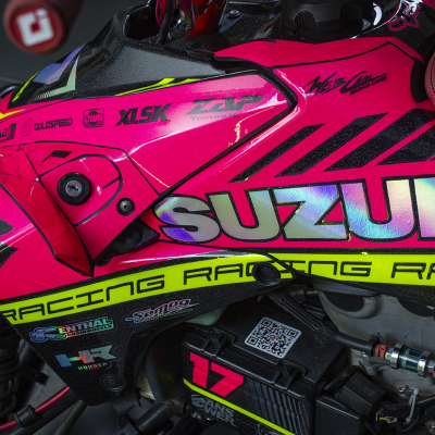 Parte lateral de mota Suzuki rosa com detalhes amarelos e pretos, banco vermelho e números 17 com QR code