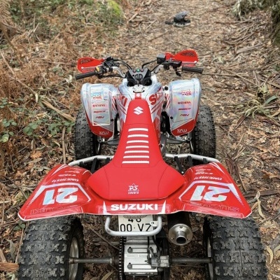 Veículo todo-o-terreno Suzuki vermelho e branco num trilho de terra com capacete preto ao fundo