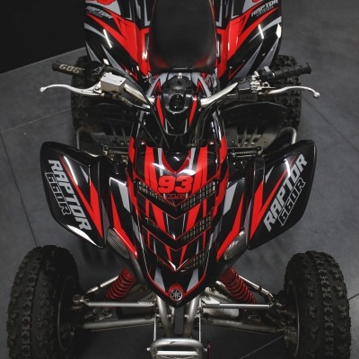 Quadriciclo vermelho e preto com adesivos Raptor 700R e 93 em fundo cinzento.
