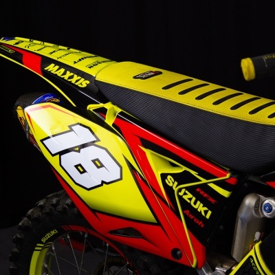 Parte traseira de mota de enduro motocross amarela, vermelha e preta com número 18 e logos MAXXIS e SUZUKI.