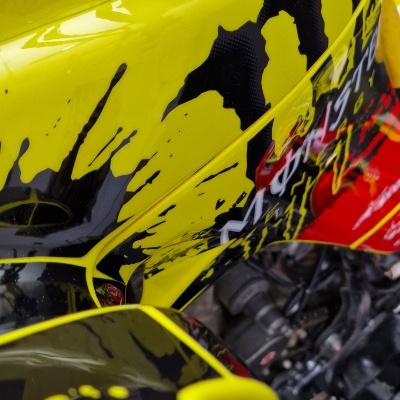 Tanque de combustível amarelo com manchas pretas e vermelhas, motor de mota ao fundo