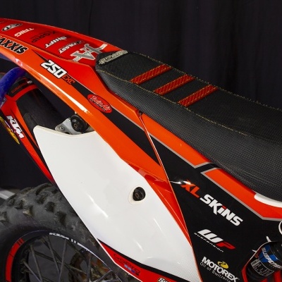 Detalhe da traseira de uma mota de motocross laranja, preto e branco com vários autocolantes
