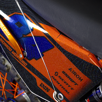 Parte lateral de mota todo-o-terreno preta com laranja e azul, suporte laranja anodizado e textos impressos.