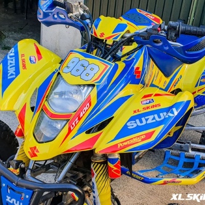 Quad Suzuki LTZ 400 amarelo, azul e vermelho com autocolantes coloridos, pneus Maxxis, em superfície de cimento com planta ao fundo