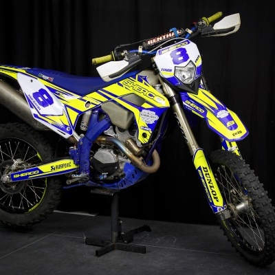 Motocross Sherco azul e amarelo com número 8 e logos visíveis em fundo preto.