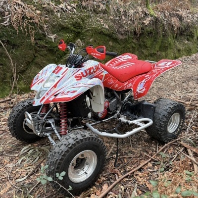 Quad Suzuki vermelho e branco com decalques vermelhos e pneus grandes todo-o-terreno num ambiente florestal