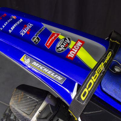Assento de moto azul com correia preta e várias etiquetas coloridas num ambiente escuro.