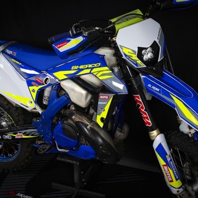 Motocross Sherco 300 SE azul, branco e amarelo com autocolantes e pneus de trilha sobre fundo negro