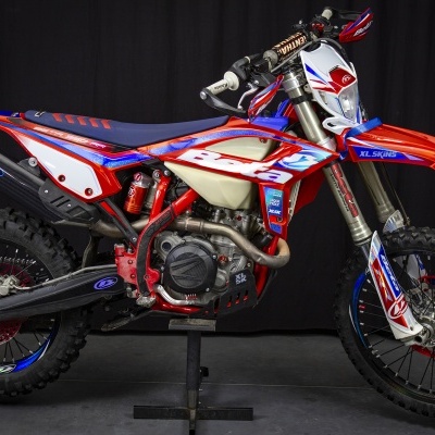 Motocross vermelho, azul e branco com pneus cravados e suporte preto.