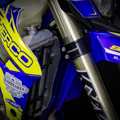 Detalhe de moto de motocross azul e amarelo com textos Sherco e Barbearia do Tiago