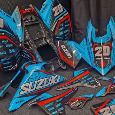 Carenagens de mota Suzuki azuis com detalhes vermelhos e pretos, texto 'SUZUKI', '20', 'Edgar Silva', várias marcas e logotipos, sobre chão cinzento