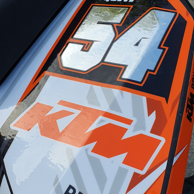 Parte lateral de uma mota KTM branca, laranja e preta com números e texto de corrida