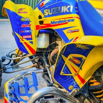 Quad Suzuki amarelo e azul com vários adesivos e proteção no guiador